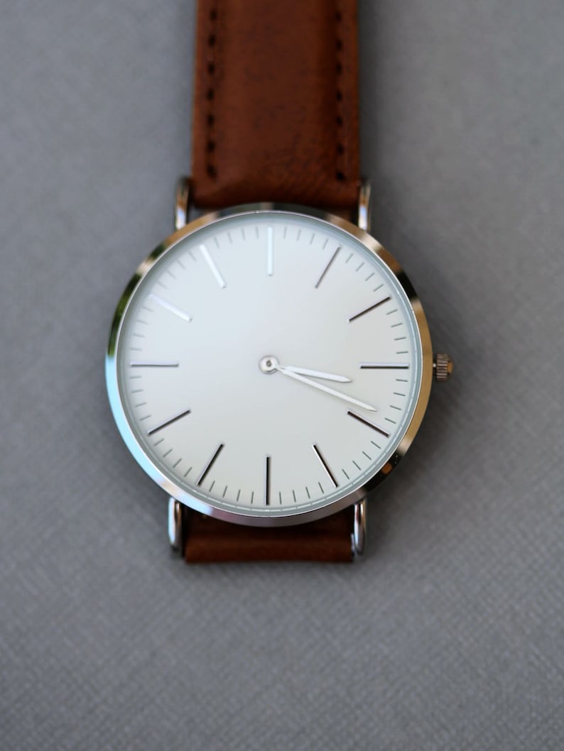Puede incluir: Un reloj plateado con una esfera blanca y una correa de cuero marr&oacute;n.
