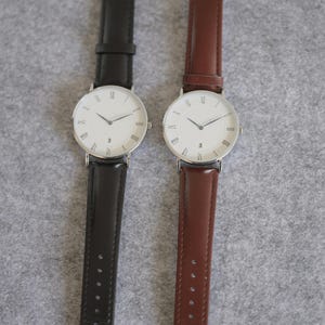 May include: Two wristwatches with white faces and Roman numeral hour markers. The watch on the left has a black leather band, and the watch on the right has a brown leather band.