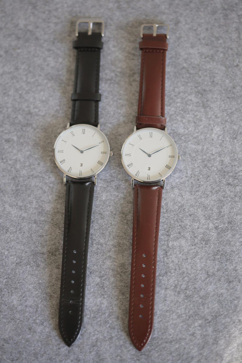 May include: Two wristwatches with white faces and Roman numeral hour markers. The watch on the left has a black leather band, and the watch on the right has a brown leather band.