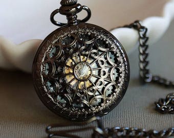 May include: A black pocket watch with a delicate, intricate design and a visible clockwork mechanism. The watch is attached to a black chain.