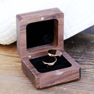 May include: A wooden ring box with a dark brown finish. The box is open and two gold rings are inside.