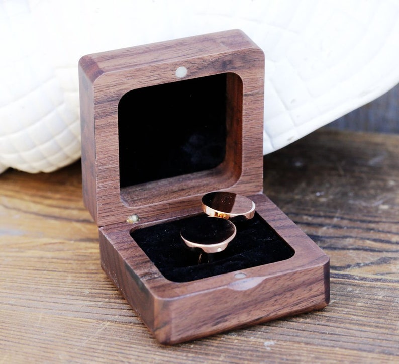 May include: A wooden ring box with a dark brown finish. The box is open and two gold rings are inside.