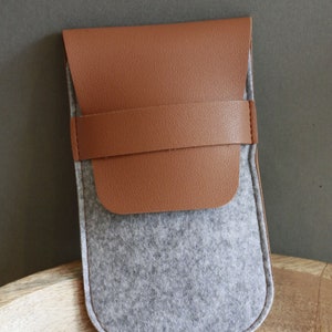 May include: A gray felt pouch with a brown leather flap and a brown leather strap. The pouch is closed and sitting on a wooden surface.