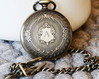May include: A gold-toned pocket watch with an ornate design and a chain. The watch face has a shield with the letter "A" engraved on it.