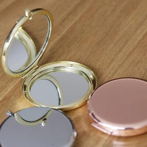 Op de afbeelding: Drie compacte spiegels, &eacute;&eacute;n goudkleurig met een scharnierend deksel, &eacute;&eacute;n goudkleurig en &eacute;&eacute;n ros&eacute; goudkleurig. De spiegels zijn rond en hebben een reflecterend oppervlak.