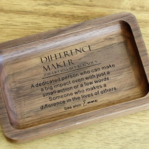 May include: A wooden tray with a definition of the word "Difference Maker" engraved on it. The definition reads: "A dedicated person who can make a big impact even with just a small action or a few words. Someone who makes a difference in the lives of others. See also: Emma."