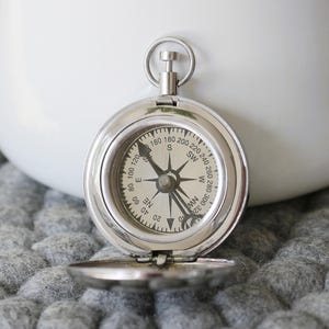May include: A silver compass with a black face and white markings. The compass is open and the needle is pointing north. The compass is sitting on a gray textured surface.