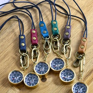 May include: Six gold compass keychains with black faces and different colored leather straps. The straps are blue, green, brown, red, and black.