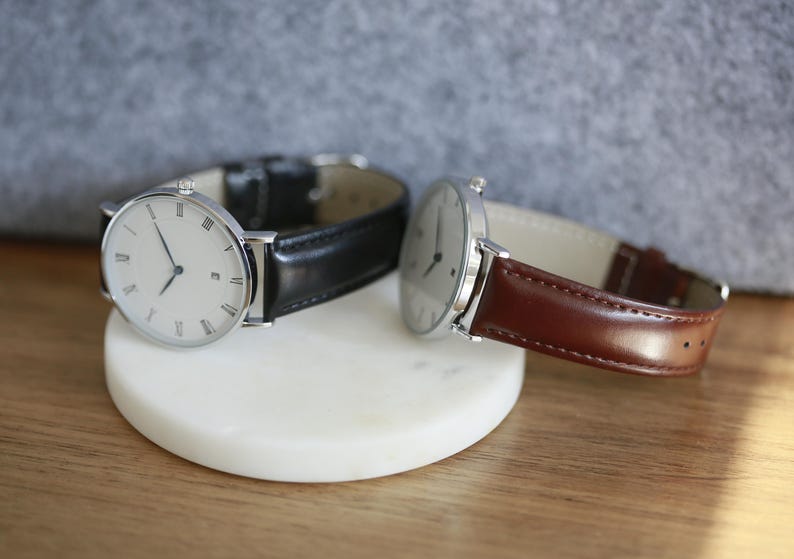 May include: Two silver wristwatches with white faces and black Roman numeral hour markers. The watch on the left has a black leather band, and the watch on the right has a brown leather band.