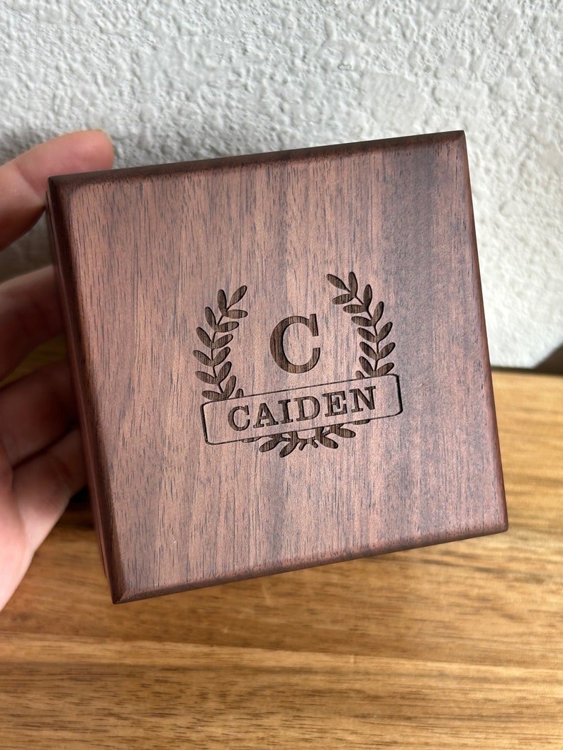 May include: A dark brown wooden box with a personalized inscription. The inscription includes a laurel wreath with the letter "C" in the center and the name "CAIDEN" below the wreath.