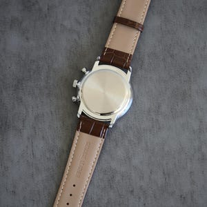 May include: A silver-toned wristwatch with a brown leather strap. The watch face is not visible, but the back of the watch and the strap are in focus. The strap is embossed with a crocodile pattern and the words "GENUINE LEATHER".