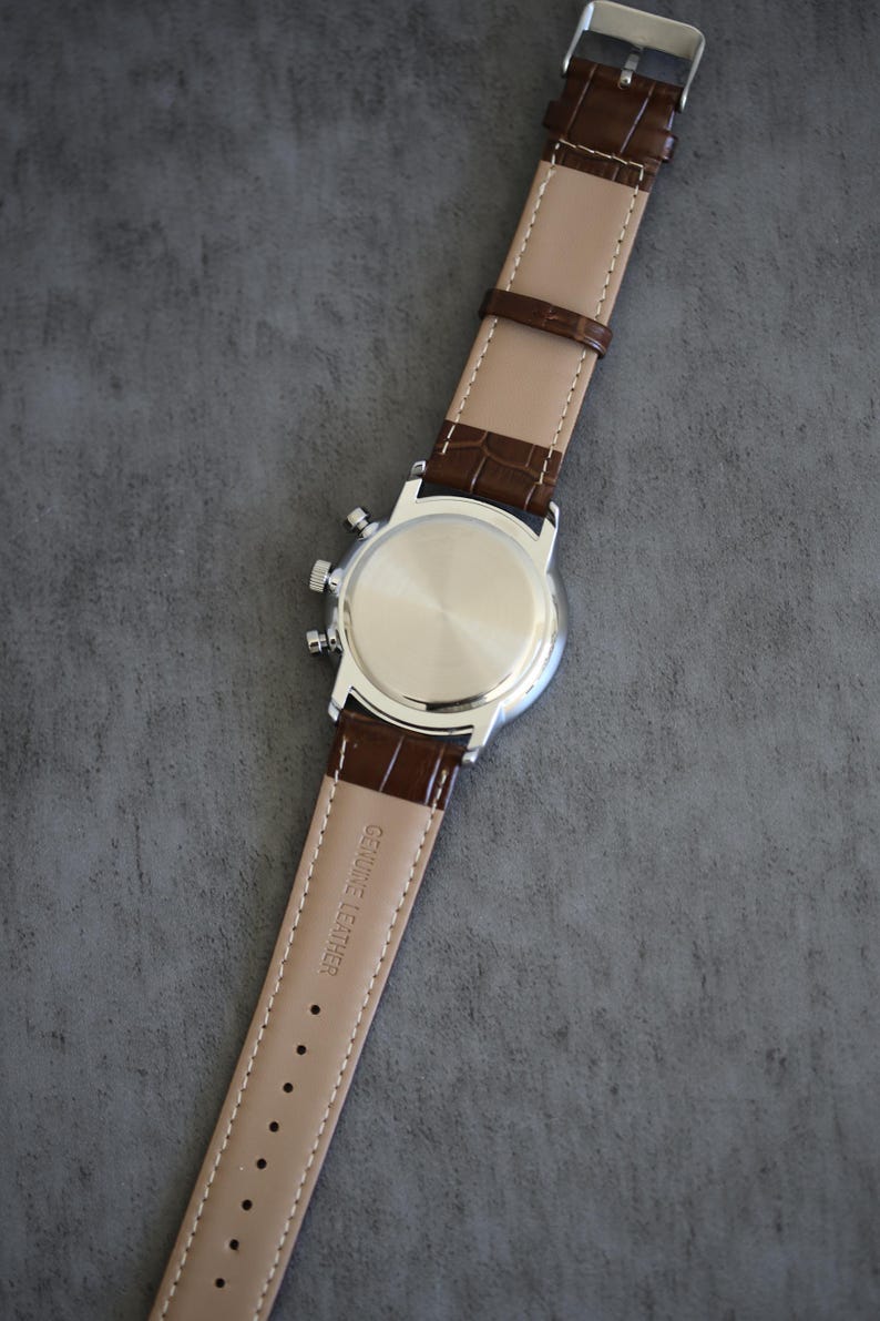 May include: A silver-toned wristwatch with a brown leather strap. The watch face is not visible, but the back of the watch and the strap are in focus. The strap is embossed with a crocodile pattern and the words "GENUINE LEATHER".
