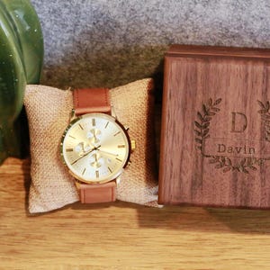 May include: A gold-toned watch with a brown leather band and a white face. The watch is displayed on a burlap cushion and comes in a wooden box with the name "Davin" engraved on it.