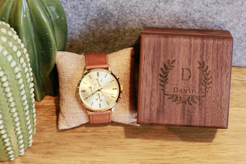 May include: A gold-toned watch with a brown leather band and a white face. The watch is displayed on a burlap cushion and comes in a wooden box with the name "Davin" engraved on it.
