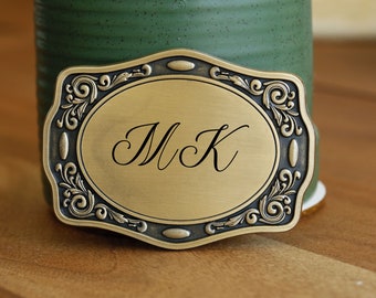 May include: A gold-toned belt buckle with an oval-shaped centre featuring the engraved initials "M K" surrounded by an ornate design.