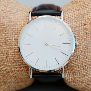 May include: A silver watch with a white face and black leather band. The watch is on a brown burlap surface.