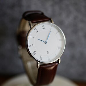 May include: A silver watch with a white face and Roman numeral hour markers. The watch has a brown leather band.