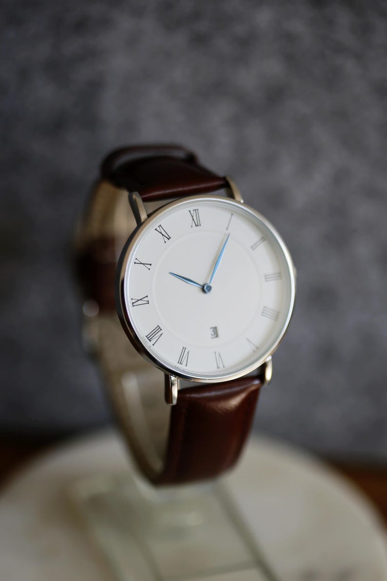 May include: A silver watch with a white face and Roman numeral hour markers. The watch has a brown leather band.