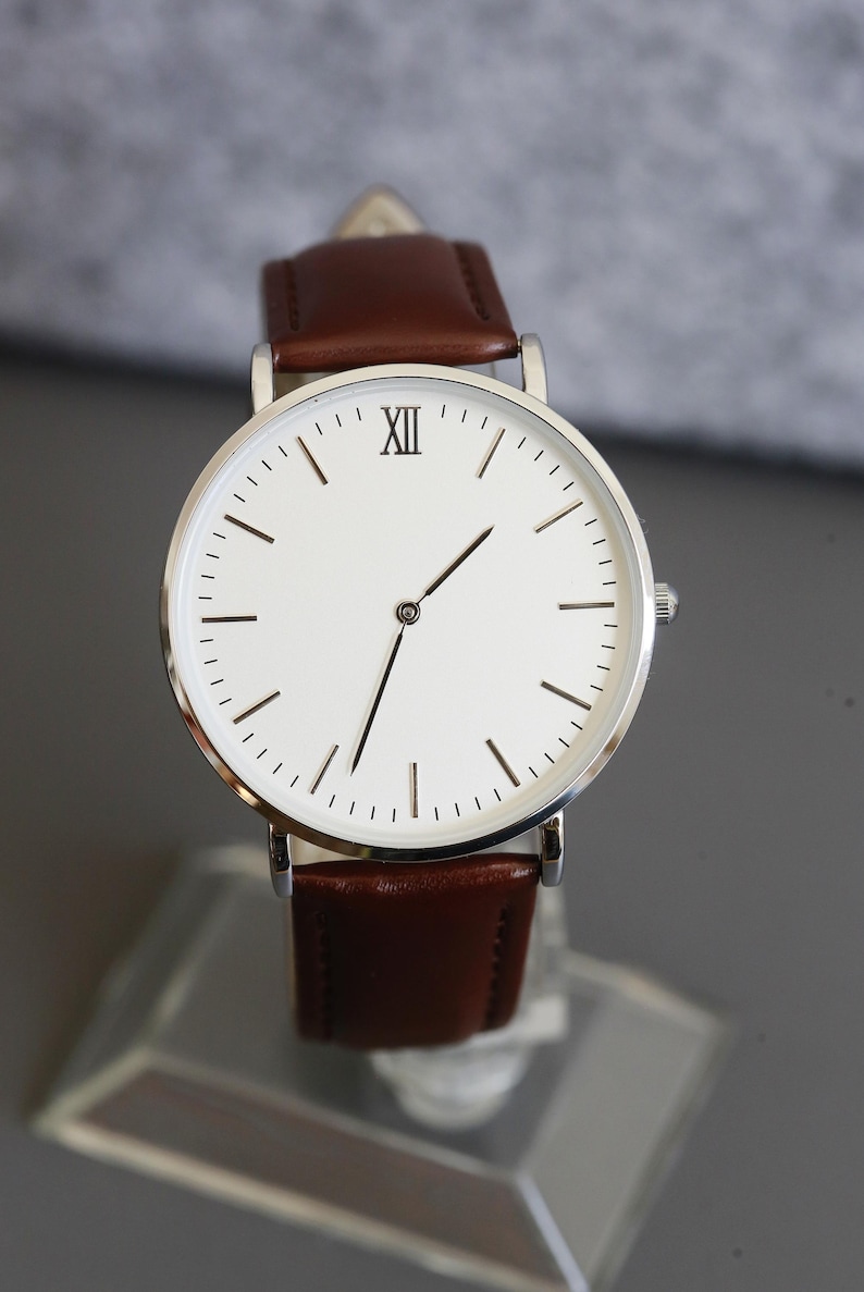 Peut inclure: Une montre argent&eacute;e avec un cadran blanc et un bracelet en cuir marron. Le cadran de la montre comporte des index en chiffres romains et une aiguille des secondes argent&eacute;e.