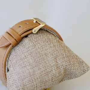 May include: A tan leather watch band with a gold buckle. The band is on a tan fabric display stand.
