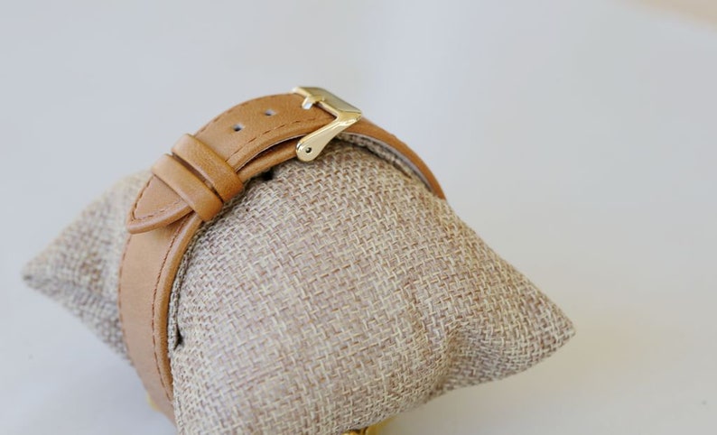 May include: A tan leather watch band with a gold buckle. The band is on a tan fabric display stand.