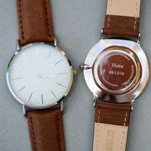 Puede incluir: Dos relojes de pulsera plateados con correas de cuero marr&oacute;n. El reloj de la izquierda tiene una esfera blanca con agujas e &iacute;ndices plateados. El reloj de la derecha tiene una parte posterior plateada con el texto "Japan Quartz Movement", "Victor", "08/12/16", "Stainless Steel Case" y "5ATM Water Resistant".