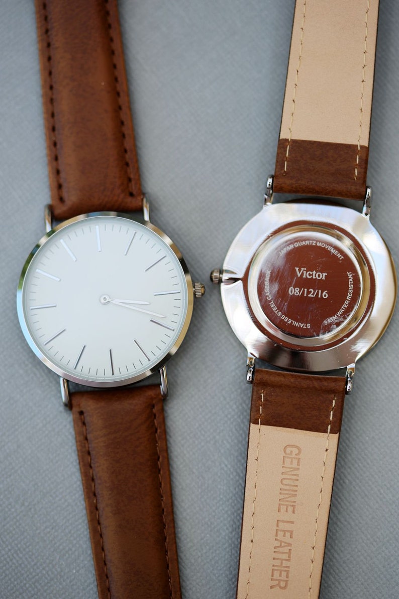 Puede incluir: Dos relojes de pulsera plateados con correas de cuero marr&oacute;n. El reloj de la izquierda tiene una esfera blanca con agujas e &iacute;ndices plateados. El reloj de la derecha tiene una parte posterior plateada con el texto "Japan Quartz Movement", "Victor", "08/12/16", "Stainless Steel Case" y "5ATM Water Resistant".