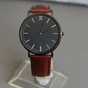 Peut inclure: Une montre noire avec un bracelet en cuir marron. Le cadran de la montre comporte des chiffres romains et des aiguilles blanches.