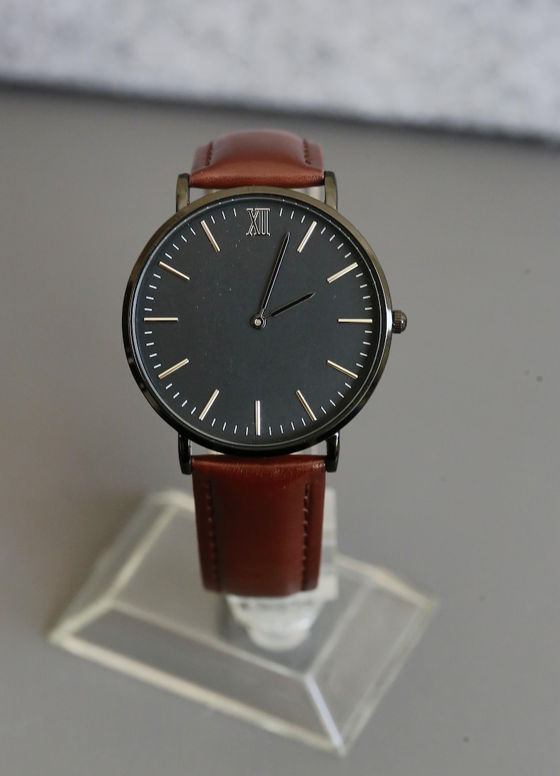 Peut inclure: Une montre noire avec un bracelet en cuir marron. Le cadran de la montre comporte des chiffres romains et des aiguilles blanches.