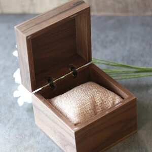 May include: A small, open wooden box with a hinged lid. The box is made of dark brown wood and contains a tan burlap pillow. The box is designed to hold a watch or other small jewelry items.
