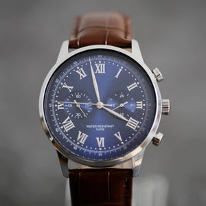 May include: A stainless steel wristwatch with a blue dial and brown leather band. The watch features Roman numeral hour markers and three sub-dials. The text "WATER RESISTANT 5 ATM" is visible on the dial. The band is a dark brown color.