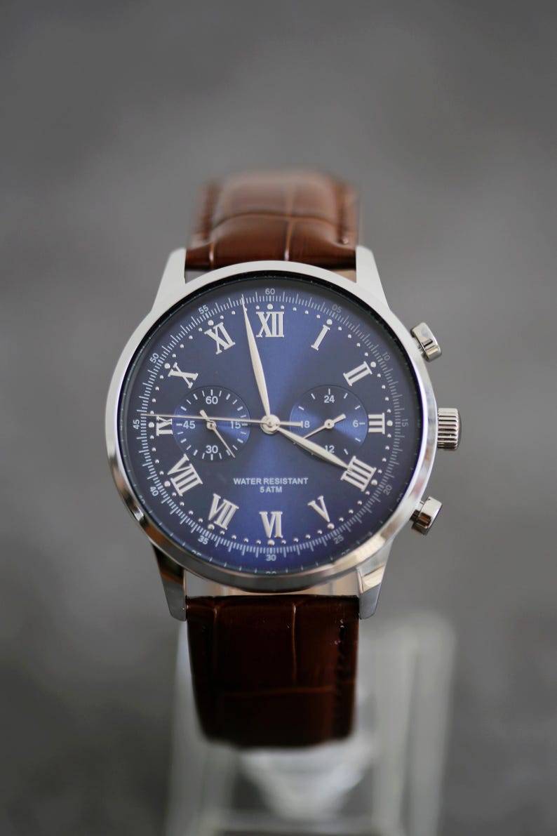 May include: A stainless steel wristwatch with a blue dial and brown leather band. The watch features Roman numeral hour markers and three sub-dials. The text "WATER RESISTANT 5 ATM" is visible on the dial. The band is a dark brown color.