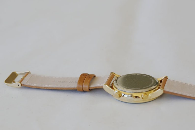 May include: A gold-tone wristwatch with a brown leather band. The watch has a round face and a gold-tone case. The band is stitched with the words "Genuine Leather".