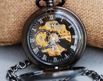 May include: A black pocket watch with a gold-toned face and Roman numeral hour markers. The watch has a skeletonized design, revealing the intricate inner workings. The watch is attached to a black chain.