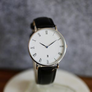 May include: A silver watch with a white face and black leather band. The watch face has Roman numeral hour markers and a blue second hand.