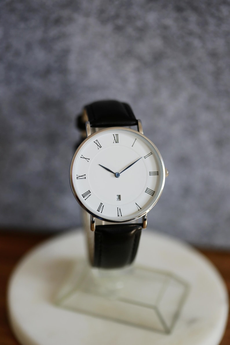 May include: A silver watch with a white face and black leather band. The watch face has Roman numeral hour markers and a blue second hand.
