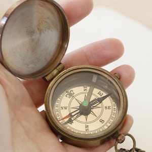 May include: A vintage brass compass with an open lid, showing a detailed compass rose. The compass has a glass cover and a chain. The compass rose features directional markings and a needle pointing north. The compass is held open.
