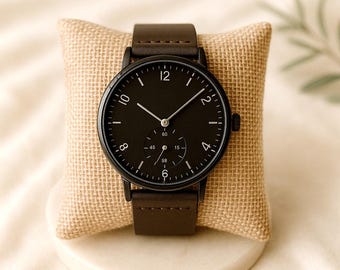Può includere: Un orologio da polso nero con cinturino in pelle marrone scuro. Il quadrante dell'orologio è nero con numeri e lancette bianche. L'orologio è esposto su un cuscino di iuta beige.
