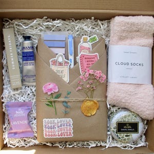 May include: A gift box filled with self-care items. Includes a hand cream, lavender sachet, stickers, a pen, a candle, a pair of pink cloud socks, and a wrapped package with a floral arrangement and a wax seal.