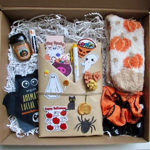 May include: A Halloween-themed gift box filled with festive items. Includes a pumpkin-scented candle, a face mask, stickers, a pen, a pair of pumpkin socks, and an orange scrunchie. The box is lined with white paper and decorated with Halloween-themed stickers.