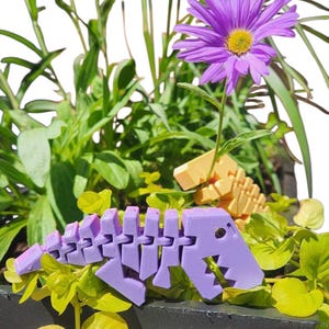 May include: A purple articulated dinosaur skeleton toy sits amongst green foliage and purple flowers in a black planter. A second, smaller, yellow dinosaur skeleton is visible. The scene is set against a white background.
