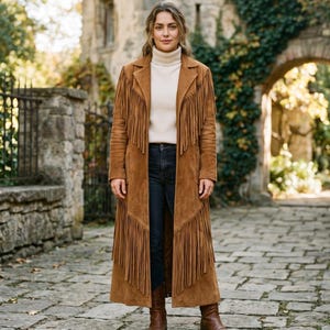 Könnte beinhalten: Ein langer, brauner Wildledermantel mit Fransen. Der Mantel wird über einem cremefarbenen Rollkragenpullover und dunkelblauen Jeans getragen. Braune Lederstiefel vervollständigen den Look. Der Mantel hat einen klassischen, böhmischen Stil.