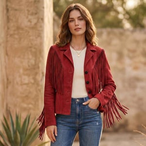 Pode incluir: Casaco de camurça vermelho com franjas longas nas mangas e no peito. O casaco tem fecho de botões e gola clássica. A modelo usa um top branco e calças de ganga azuis.