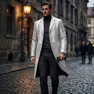 May include: A silver, long leather coat with a crocodile skin texture. The coat is worn over a dark gray turtleneck sweater and dark gray pants. The person is holding brown gloves. The background is a cobblestone street.