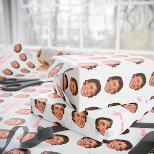 May include: White wrapping paper with a repeating pattern of smiling faces. Two wrapped gift boxes are stacked, with a roll of paper and a sheet of paper with the same face pattern. A pair of scissors and a black ribbon are also visible.
