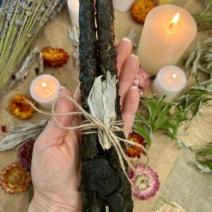 Może przedstawiać: Dwie ciemnozielone świece rytualne związane sznurkiem, z czarną pieczęcią woskową z motywem słońca. Świece otoczone są suszonymi kwiatami, szałwią i zapalonymi świecami, tworząc mistyczną i duchową estetykę.