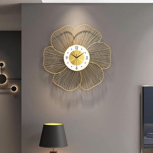 Könnte beinhalten: Eine goldfarbene Wanduhr aus Metall in Form einer Blume. Das Zifferblatt der Uhr ist weiß mit schwarzen Zahlen und Zeigern. Die Blütenblätter bestehen aus dünnen, gebogenen Metalllinien. Die Uhr ist an einer grauen Wand befestigt.