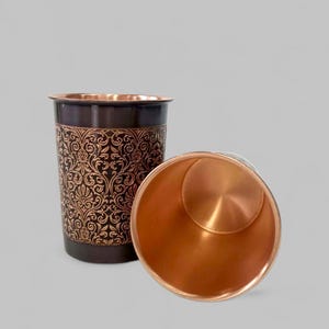 Puede incluir: Dos vasos de metal color cobre. Un vaso está en posición vertical y el otro está tumbado de lado. El vaso vertical tiene una banda marrón oscuro en la parte superior y un patrón floral decorativo. El interior de los vasos es de color cobre brillante.