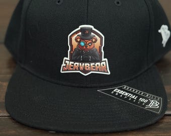 Pode incluir: Boné snapback preto com um patch de urso usando cartola e monóculo, com o texto "JERVBEAR". O boné tem uma aba reta e uma etiqueta "Branded Bills".