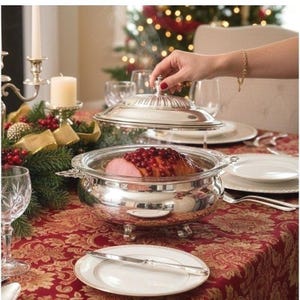 Puede incluir: Un plato de servicio de plata pulida con tapa a juego, que presenta asas ornamentadas y cuatro patas. El recipiente redondo contiene un jamón cocido cubierto con arándanos rojos, con vapor saliendo del plato al levantar la tapa de la base.